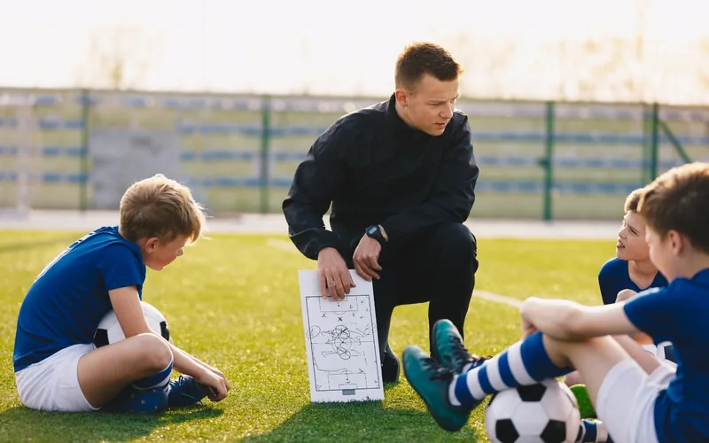 Soccer coach explaining tactics with a board to young players sitting on the field